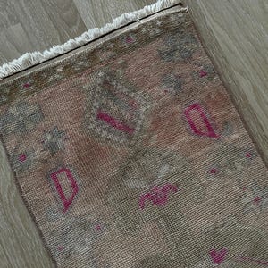 May include: A rectangular, distressed rug with a faded, vintage aesthetic. The rug features a blend of muted pink, gray, and brown tones, with pops of bright pink geometric shapes. The edges are finished with a fringe.