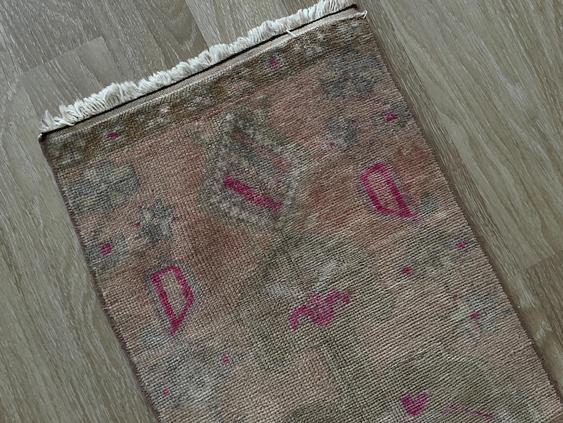 May include: A rectangular, distressed rug with a faded, vintage aesthetic. The rug features a blend of muted pink, gray, and brown tones, with pops of bright pink geometric shapes. The edges are finished with a fringe.