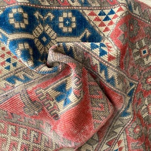 May include: A woven rug featuring a geometric pattern in shades of red, blue, and beige. The textile displays intricate diamond and floral motifs with a textured pile, showcasing a folded corner that reveals the detailed construction of the fabric.