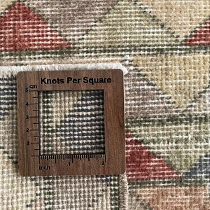 K&ouml;nnte beinhalten: Nahaufnahme eines Vintage-Teppichs mit geometrischem Muster in Beige-, Rot- und Blaut&ouml;nen. Ein Holzwerkzeug mit dem Text "Knots Per Square" und Zoll- und cm-Ma&szlig;en liegt auf dem Teppich.