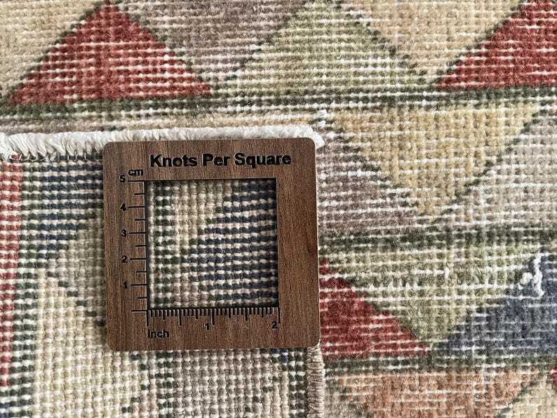 K&ouml;nnte beinhalten: Nahaufnahme eines Vintage-Teppichs mit geometrischem Muster in Beige-, Rot- und Blaut&ouml;nen. Ein Holzwerkzeug mit dem Text "Knots Per Square" und Zoll- und cm-Ma&szlig;en liegt auf dem Teppich.