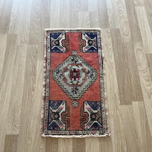 Puede incluir: Una pequeña alfombra rectangular con un patrón geométrico. La alfombra presenta una forma de diamante central en gris y rojo, rodeada de detalles azules y rojos. La alfombra tiene un borde con flecos y está colocada sobre un suelo de madera.
