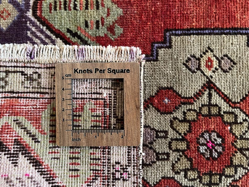 May include: Close-up of a section of a vintage rug with a wooden ruler that reads "Knots Per Square" in the image. The rug features a complex geometric pattern in red, green, purple, and beige. The ruler shows measurements in inches and centimeters.
