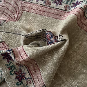 Puede incluir: Primer plano de una alfombra vintage beige y rosa con intrincados patrones florales y geométricos. La alfombra tiene un aspecto desgastado, con colores descoloridos y desgaste visible. Los bordes presentan un borde con diseños detallados.