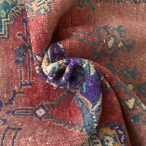 Puede incluir: Una alfombra tejida con un color base rojo intenso y patrones geométricos y florales intrincados en tonos verde azulado, morado y crema. La tela muestra un pelo texturizado con una esquina doblada que revela la estructura del tejido.