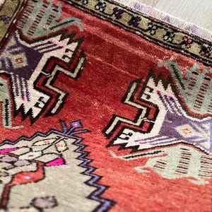 May include: Close-up of a small, rectangular rug with a distressed, vintage appearance. The rug features a central field of red with geometric patterns in white, purple, and green. The border is a darker green with floral motifs. Fringes are visible.