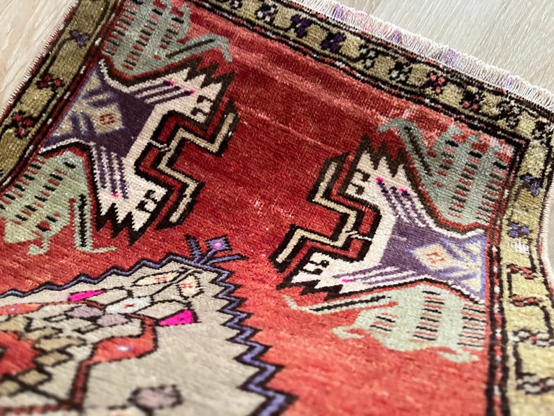 May include: Close-up of a small, rectangular rug with a distressed, vintage appearance. The rug features a central field of red with geometric patterns in white, purple, and green. The border is a darker green with floral motifs. Fringes are visible.