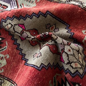 May include: Close-up of a vintage rug with a distressed, textured appearance. The rug features a red background with geometric patterns in beige, purple, and green. The design includes zig-zag borders and floral motifs, showcasing a traditional style.