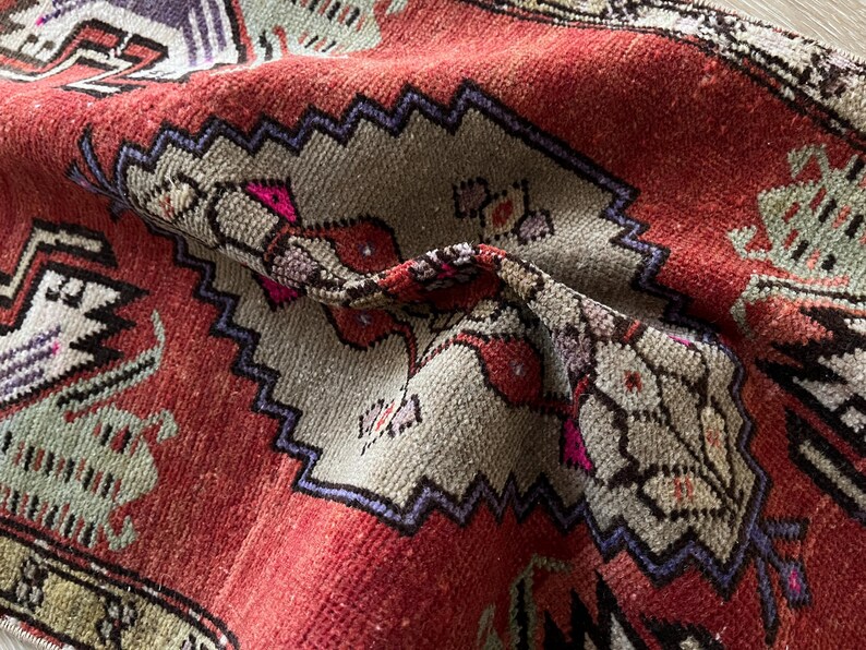 May include: Close-up of a vintage rug with a distressed, textured appearance. The rug features a red background with geometric patterns in beige, purple, and green. The design includes zig-zag borders and floral motifs, showcasing a traditional style.