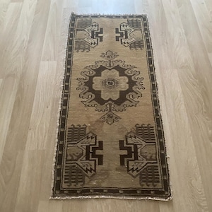 May include: A rectangular runner rug featuring a tan background with dark brown geometric patterns. The design includes a central floral-inspired medallion flanked by symmetrical angular motifs at both ends, finished with a thin dark border.
