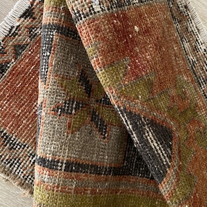 May include: A woven rug featuring a geometric pattern in shades of rust orange, olive green, and charcoal black. The textile has a visible pile texture and is finished with short, light-colored fringe along the edges, showing a folded corner.