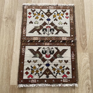 Pode incluir: Uma tapeçaria retangular de tecido entrelaçado com dois painéis simétricos sobre um fundo creme. Cada painel exibe motivos de pássaros em castanho, elementos florais em amarelo, vermelho e azul, e pequenos corações, com uma borda castanha.