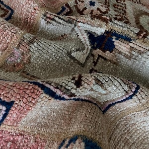 May include: Close-up of a vintage rug with a distressed, textured appearance. The rug features a mix of colors, including beige, pink, brown, and navy blue. The design includes geometric and floral patterns, creating a traditional aesthetic.