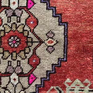 May include: Close-up of a section of a hand-knotted rug. The rug features a central design in shades of red, pink, and beige, with a border in a darker red and blue. The rug's texture is visible, showcasing the intricate weaving.