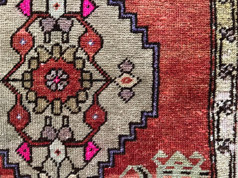 May include: Close-up of a section of a hand-knotted rug. The rug features a central design in shades of red, pink, and beige, with a border in a darker red and blue. The rug's texture is visible, showcasing the intricate weaving.