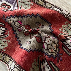 May include: Close-up of a vintage red and beige rug with intricate geometric patterns. The rug features a central design with floral and geometric motifs, framed by a border with repeating shapes. The colors include red, beige, purple, and green.