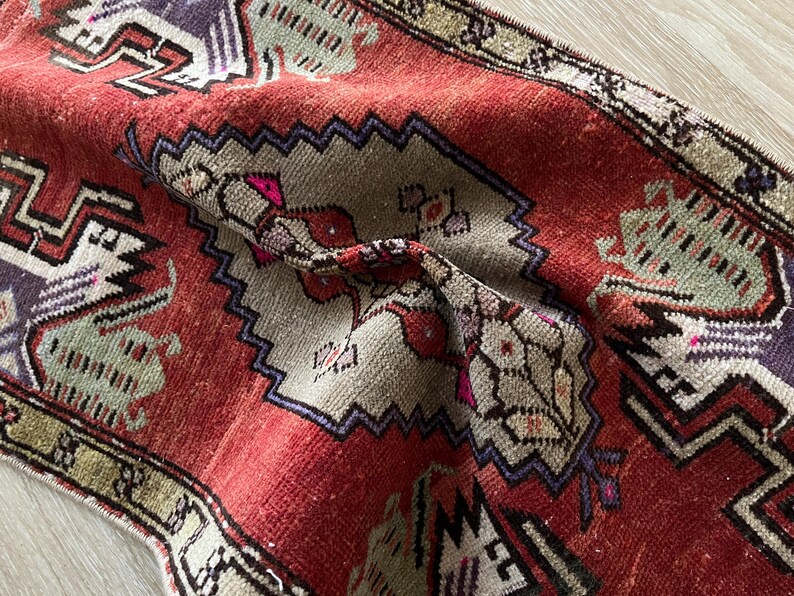May include: Close-up of a vintage red and beige rug with intricate geometric patterns. The rug features a central design with floral and geometric motifs, framed by a border with repeating shapes. The colors include red, beige, purple, and green.