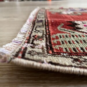 May include: Close-up of a small, rectangular rug with a detailed pattern. The rug features a red field with intricate designs in green, black, and purple. The border is beige with a repeating geometric pattern. Fringes are visible on the edge.