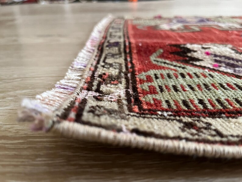 May include: Close-up of a small, rectangular rug with a detailed pattern. The rug features a red field with intricate designs in green, black, and purple. The border is beige with a repeating geometric pattern. Fringes are visible on the edge.