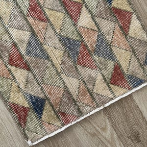 K&ouml;nnte beinhalten: Ein rechteckiger Teppich mit einem geometrischen Muster aus Dreiecken in verschiedenen Farben, darunter Rot, Blau, Beige und Gr&uuml;n. Der Teppich hat einen Used-Look im Vintage-Stil mit einem wei&szlig;en Fransenrand. Er liegt auf einem Holzboden.