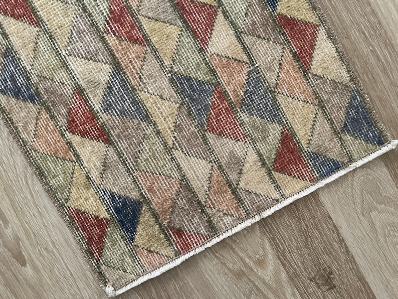 K&ouml;nnte beinhalten: Ein rechteckiger Teppich mit einem geometrischen Muster aus Dreiecken in verschiedenen Farben, darunter Rot, Blau, Beige und Gr&uuml;n. Der Teppich hat einen Used-Look im Vintage-Stil mit einem wei&szlig;en Fransenrand. Er liegt auf einem Holzboden.