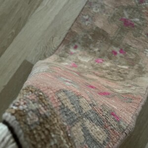 May include: A rolled-up, rectangular rug with a faded, vintage appearance. The rug features a blend of muted colors, including pink, gray, and brown, with small, bright pink accents. The rug has a fringed edge.