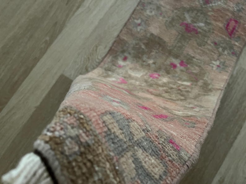 May include: A rolled-up, rectangular rug with a faded, vintage appearance. The rug features a blend of muted colors, including pink, gray, and brown, with small, bright pink accents. The rug has a fringed edge.