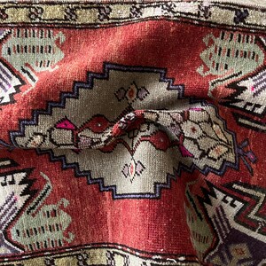 May include: Close-up of a folded, vintage rug with a red central field and intricate geometric patterns. The rug features a central medallion with a stepped border in shades of gray, purple, and green. The edges have a decorative border.