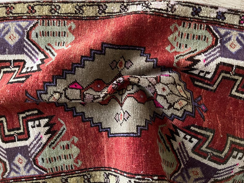 May include: Close-up of a folded, vintage rug with a red central field and intricate geometric patterns. The rug features a central medallion with a stepped border in shades of gray, purple, and green. The edges have a decorative border.