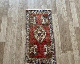 Puede incluir: Una alfombra rectangular tejida a mano con un campo central rojo y patrones geométricos intrincados en marrón, beige y crema. La alfombra presenta un medallón central y motivos de animales estilizados, con un borde con flecos. Mide aproximadamente 45 x 91 cm.