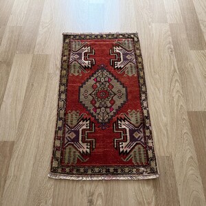 May include: A rectangular, hand-woven rug with a red background and intricate geometric patterns in shades of beige, purple, and green. The rug has a central medallion design and a detailed border. The rug is approximately 24 inches long and 16 inches wide.