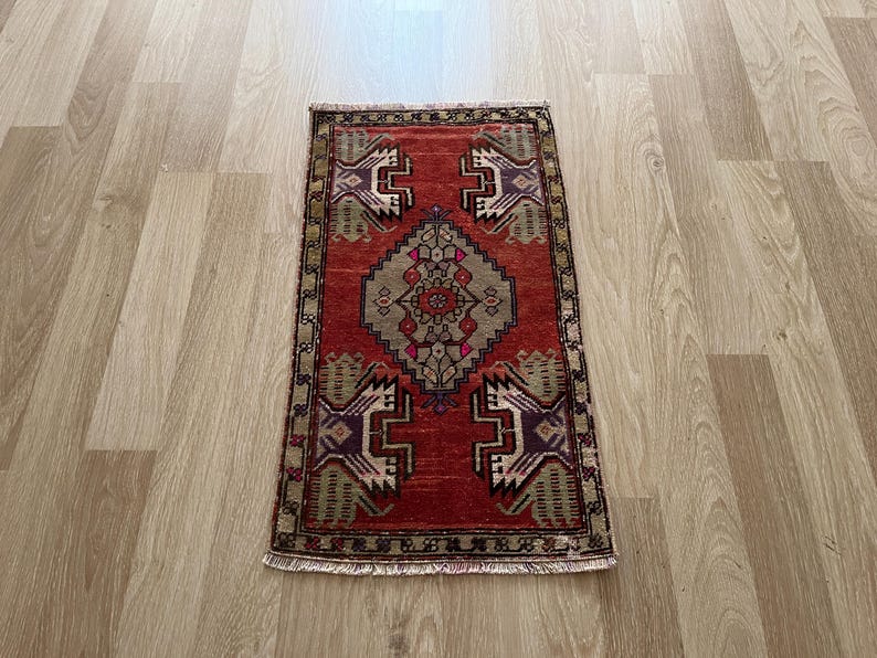 May include: A rectangular, hand-woven rug with a red background and intricate geometric patterns in shades of beige, purple, and green. The rug has a central medallion design and a detailed border. The rug is approximately 24 inches long and 16 inches wide.