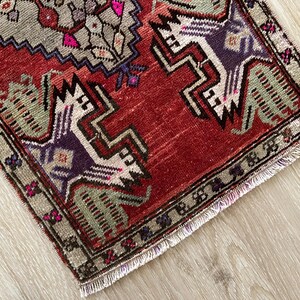 May include: A small, rectangular rug with a distressed, vintage appearance. The rug features a red field with geometric designs in shades of gray, purple, and green. The border is a muted green with small floral accents. The rug has a fringed edge.