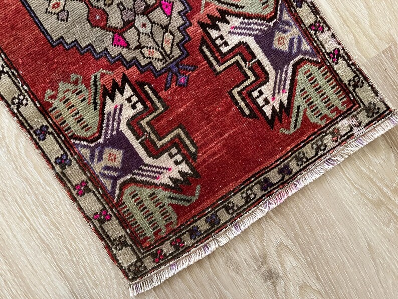 May include: A small, rectangular rug with a distressed, vintage appearance. The rug features a red field with geometric designs in shades of gray, purple, and green. The border is a muted green with small floral accents. The rug has a fringed edge.