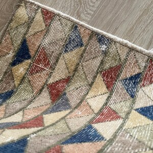 K&ouml;nnte beinhalten: Kleiner, rechteckiger Teppich mit einem geometrischen Muster aus Dreiecken in Beige-, Rot-, Blau- und Gelbt&ouml;nen. Der Teppich hat einen Used-Look im Vintage-Stil mit einem Fransenrand. Das Design zeigt eine Reihe von gebogenen Linien.