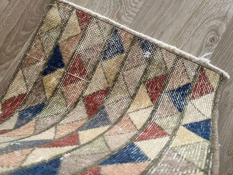 K&ouml;nnte beinhalten: Kleiner, rechteckiger Teppich mit einem geometrischen Muster aus Dreiecken in Beige-, Rot-, Blau- und Gelbt&ouml;nen. Der Teppich hat einen Used-Look im Vintage-Stil mit einem Fransenrand. Das Design zeigt eine Reihe von gebogenen Linien.