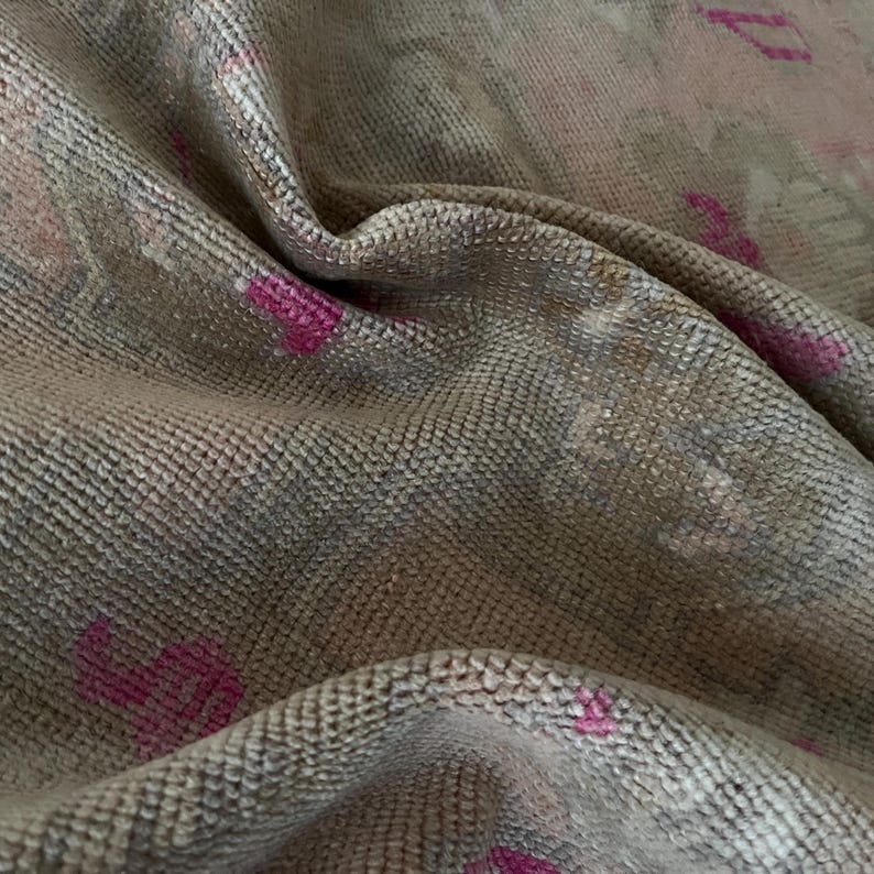 May include: Close-up of a textured, woven fabric with a neutral color palette. The fabric has a subtle pattern and is accented with small, bright pink floral or heart-shaped designs. The material is draped, showing folds and shadows.