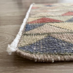 K&ouml;nnte beinhalten: Nahaufnahme eines kleinen, rechteckigen Teppichs mit geometrischem Muster. Der Teppich weist eine Mischung aus Farben auf, darunter Rot, Blau, Beige und Gr&uuml;n. Die Kanten sind mit einem Fransen versehen.