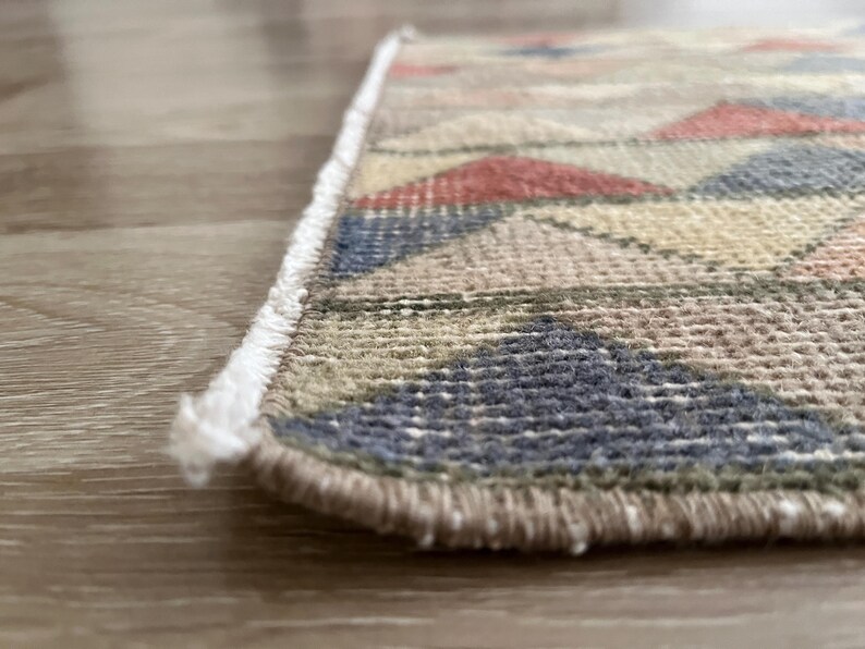 K&ouml;nnte beinhalten: Nahaufnahme eines kleinen, rechteckigen Teppichs mit geometrischem Muster. Der Teppich weist eine Mischung aus Farben auf, darunter Rot, Blau, Beige und Gr&uuml;n. Die Kanten sind mit einem Fransen versehen.