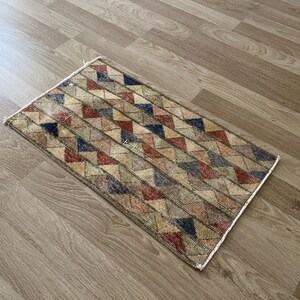 K&ouml;nnte beinhalten: Rechteckiger Teppich mit einem geometrischen Muster aus roten, blauen, beigen und gr&uuml;nen Dreiecken. Der Teppich hat ein verwittertes, Vintage-Aussehen und liegt auf einem hellbraunen Holzboden.