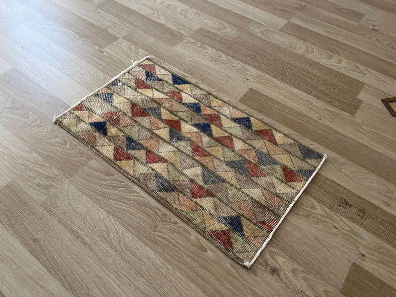 K&ouml;nnte beinhalten: Rechteckiger Teppich mit einem geometrischen Muster aus roten, blauen, beigen und gr&uuml;nen Dreiecken. Der Teppich hat ein verwittertes, Vintage-Aussehen und liegt auf einem hellbraunen Holzboden.