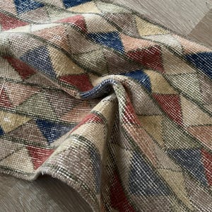 K&ouml;nnte beinhalten: Eine aufgerollte, Vintage-Teppich mit einem geometrischen Muster aus Dreiecken in Beige, Rot, Blau und Gr&uuml;n. Der Teppich hat einen Used-Look mit sichtbaren Abnutzungserscheinungen, was seinen antiken Charme unterstreicht. Die Textur des Teppichs wirkt weich und gewebt.