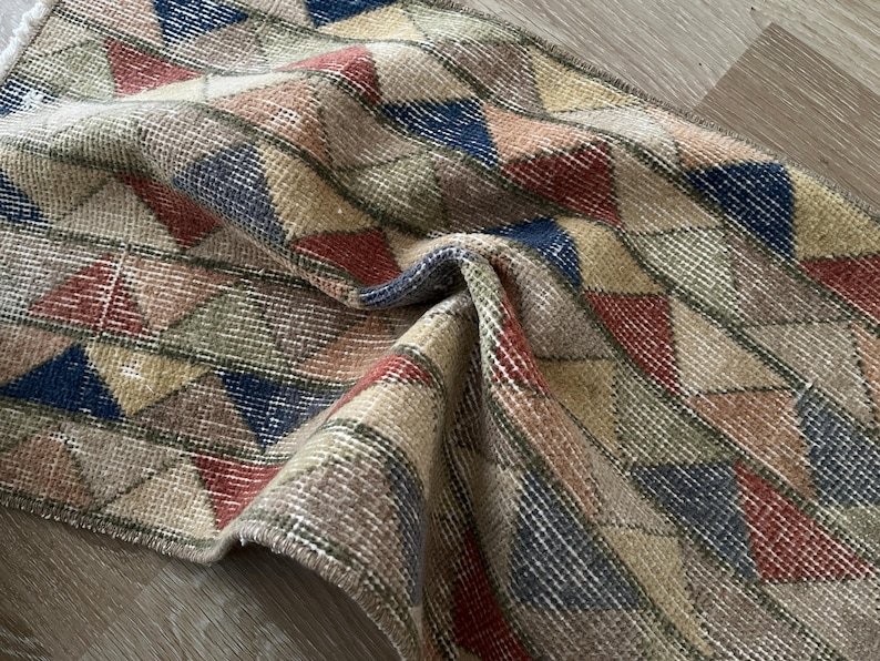 K&ouml;nnte beinhalten: Eine aufgerollte, Vintage-Teppich mit einem geometrischen Muster aus Dreiecken in Beige, Rot, Blau und Gr&uuml;n. Der Teppich hat einen Used-Look mit sichtbaren Abnutzungserscheinungen, was seinen antiken Charme unterstreicht. Die Textur des Teppichs wirkt weich und gewebt.
