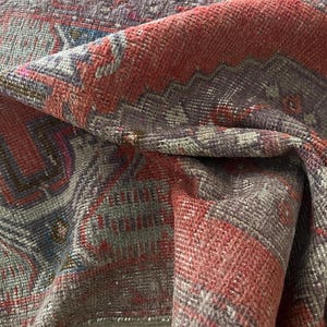 May include: Close-up of a folded, vintage rug with a distressed appearance. The rug features a complex pattern in shades of red, gray, and purple. The design includes geometric shapes and floral motifs, creating a textured, antique aesthetic.