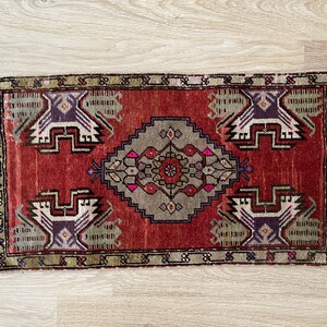 May include: A rectangular, hand-woven rug with a central diamond pattern in gray, pink, and brown. The rug features a red background with repeating geometric designs in white, purple, and green. The edges have a fringed border.