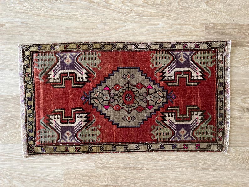 May include: A rectangular, hand-woven rug with a central diamond pattern in gray, pink, and brown. The rug features a red background with repeating geometric designs in white, purple, and green. The edges have a fringed border.