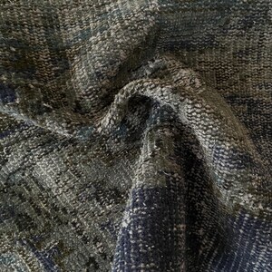 May include: Close-up of a textured rug with a mix of dark blue, green, and gray tones. The rug's surface has a woven appearance with visible variations in color and texture, creating a vintage or distressed look.