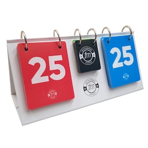 Puede incluir: Un marcador de mesa de metal blanco con tres tarjetas abatibles. Las tarjetas son rojas, negras y azules, cada una con el número 25 en blanco. El marcador tiene un logotipo en las tarjetas y la base.