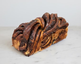 May include: A loaf of freshly baked babka, a sweet braided bread. The bread has a rich, dark brown color with visible layers of chocolate filling. The babka sits on a white marble surface, showcasing its texture and form.