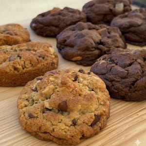 May include: An assortment of freshly baked cookies, including chocolate chip and double chocolate varieties, arranged on a light wooden surface. Some cookies have visible nuts and caramel pieces, showcasing a variety of textures and flavors.